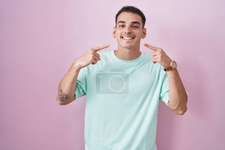 Téléchargez les photos : Bel homme hispanique debout sur fond rose souriant gai montrant et pointant avec les doigts dents et la bouche. concept de santé dentaire. - en image libre de droit