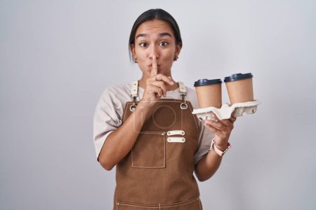 Photo for Young hispanic woman wearing professional waitress apron holding coffee asking to be quiet with finger on lips. silence and secret concept. - Royalty Free Image