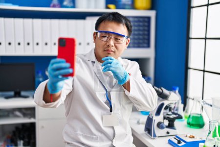 Foto de Young chinese man wearing scientist uniform having video call at laboratory - Imagen libre de derechos