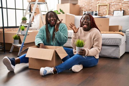 Téléchargez les photos : Deux femmes africaines déménageant dans une nouvelle maison assise sur le sol célébrant la victoire avec un sourire heureux et l'expression gagnante avec les mains levées - en image libre de droit