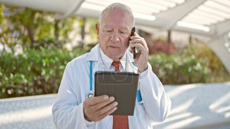 Foto de Senior médico hombre de pelo gris usando touchpad hablando en el teléfono inteligente en el parque - Imagen libre de derechos
