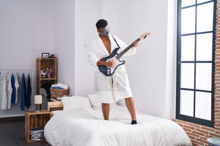 Téléchargez les photos : Jeune homme latin jouant de la guitare électrique sur le lit avec masque facial traitement dans la chambre - en image libre de droit