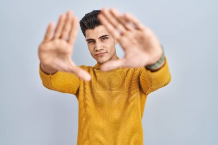 Foto de Joven hombre hispano de pie sobre fondo azul haciendo marco usando manos palmas y dedos, perspectiva de la cámara - Imagen libre de derechos