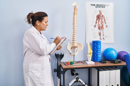 Photo for Middle age hispanic woman physiotherapist pointing to anatomical model of vertebral column at rehab clinic - Royalty Free Image