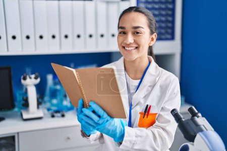 Photo for Young hispanic woman scientist reading book at laboratory - Royalty Free Image
