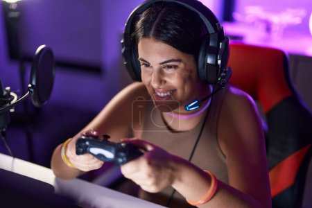 Foto de Young beautiful hispanic woman streamer playing video game using joystick at gaming room - Imagen libre de derechos