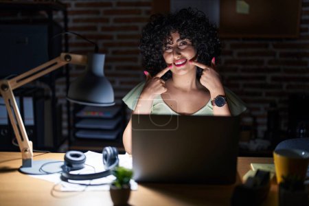 Photo for Young brunette woman with curly hair working at the office at night smiling cheerful showing and pointing with fingers teeth and mouth. dental health concept. - Royalty Free Image