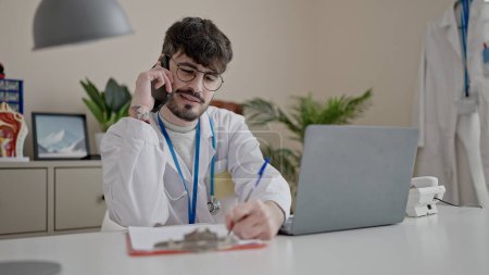 Photo for Young hispanic man doctor talking on telephone writing on document at clinic - Royalty Free Image