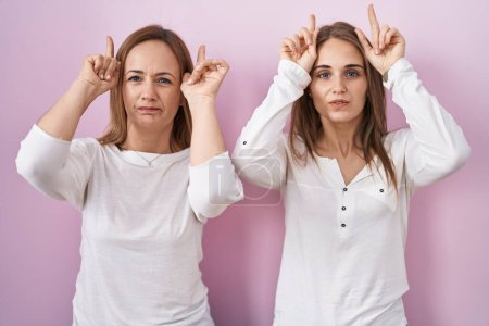 Photo for Middle age mother and young daughter standing over pink background doing funny gesture with finger over head as bull horns - Royalty Free Image