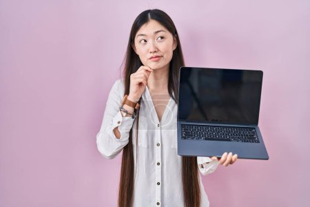 Foto de Mujer joven china sosteniendo el ordenador portátil que muestra la cara seria de la pantalla pensando en la pregunta con la mano en la barbilla, reflexivo sobre la idea confusa - Imagen libre de derechos