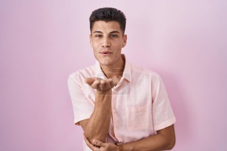 Téléchargez les photos : Jeune homme hispanique debout sur fond rose regardant la caméra souffler un baiser avec la main sur l'air étant belle et sexy. expression d'amour. - en image libre de droit
