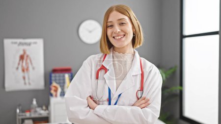 Foto de Joven rubia doctora sonriendo confiada de pie con los brazos cruzados gesto en la clínica - Imagen libre de derechos