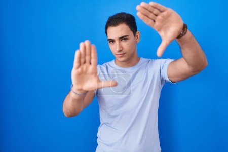 Photo for Young hispanic man standing over blue background doing frame using hands palms and fingers, camera perspective - Royalty Free Image