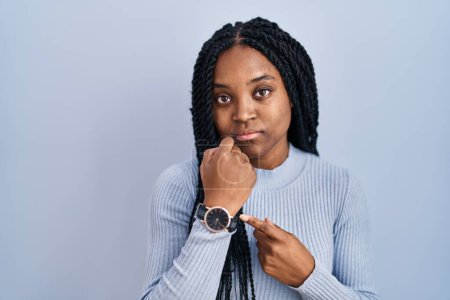 Téléchargez les photos : Femme afro-américaine debout sur fond bleu pressé pointant vers regarder le temps, l'impatience, regardant la caméra avec une expression détendue - en image libre de droit