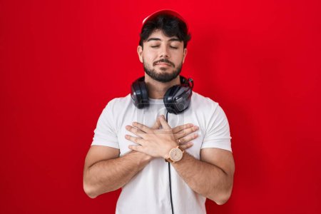 Photo for Hispanic man with beard wearing gamer hat and headphones smiling with hands on chest with closed eyes and grateful gesture on face. health concept. - Royalty Free Image