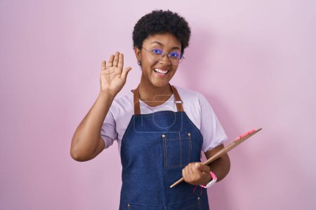 Photo for Young african american woman wearing professional waitress apron holding clipboard waiving saying hello happy and smiling, friendly welcome gesture - Royalty Free Image