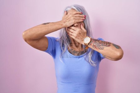 Foto de Mujer de mediana edad con tatuajes de pie sobre fondo rosa que cubre los ojos y la boca con las manos, sorprendida y sorprendida. ocultar emociones - Imagen libre de derechos