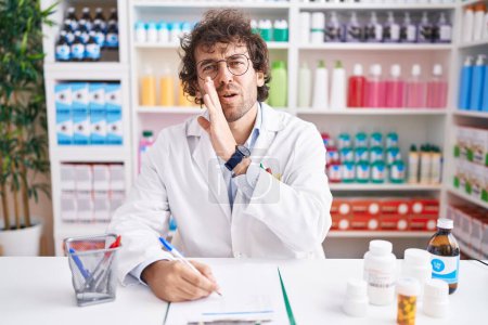 Photo for Hispanic young man working at pharmacy drugstore hand on mouth telling secret rumor, whispering malicious talk conversation - Royalty Free Image