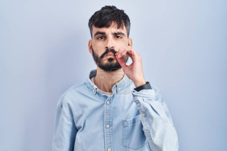 Photo for Young hispanic man with beard standing over blue background mouth and lips shut as zip with fingers. secret and silent, taboo talking - Royalty Free Image