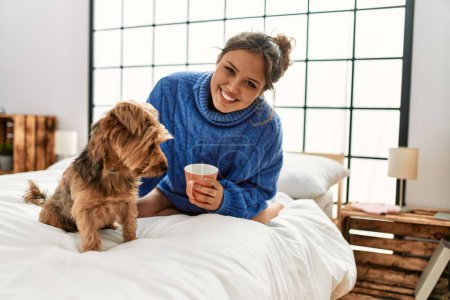Foto de Joven mujer hispana hermosa bebiendo taza de café sentado en la cama con perro en el dormitorio - Imagen libre de derechos