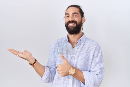 Foto de Hombre hispano con barba vistiendo camisa casual mostrando la palma de la mano y haciendo buen gesto con los pulgares hacia arriba, sonriendo feliz y alegre - Imagen libre de derechos