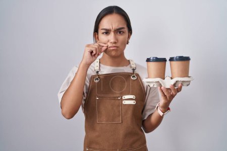 Téléchargez les photos : Jeune femme hispanique portant tablier de serveuse professionnel tenant bouche de café et lèvres fermées comme zip avec les doigts. secret et silencieux, tabou parlant - en image libre de droit