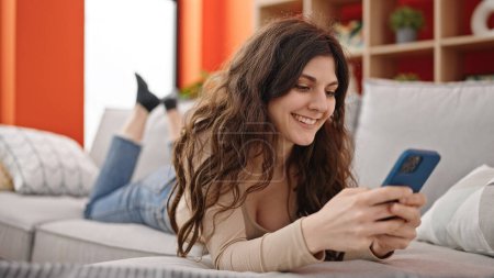 Photo for Young beautiful hispanic woman using smartphone lying on sofa at home - Royalty Free Image