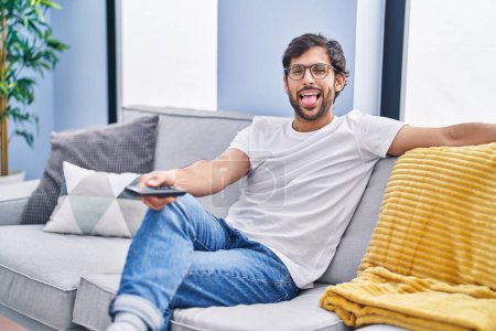 Foto de Hombre latino guapo sosteniendo el control remoto de la televisión sacando la lengua feliz con expresión divertida. - Imagen libre de derechos