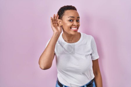 Foto de Hermosa mujer afroamericana de pie sobre fondo rosa sonriendo con la mano sobre el oído escuchando un rumor o chismes. concepto de sordera. - Imagen libre de derechos