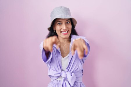 Téléchargez les photos : Jeune femme hispanique debout sur fond rose portant un chapeau pointant vers vous et la caméra avec les doigts, souriant positif et joyeux - en image libre de droit