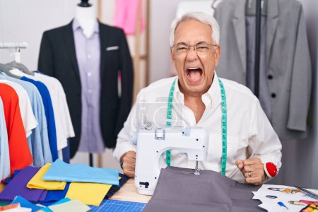 Photo for Middle age man with grey hair dressmaker using sewing machine angry and mad screaming frustrated and furious, shouting with anger. rage and aggressive concept. - Royalty Free Image