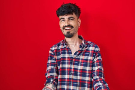 Photo for Young hispanic man with beard standing over red background smiling cheerful with open arms as friendly welcome, positive and confident greetings - Royalty Free Image
