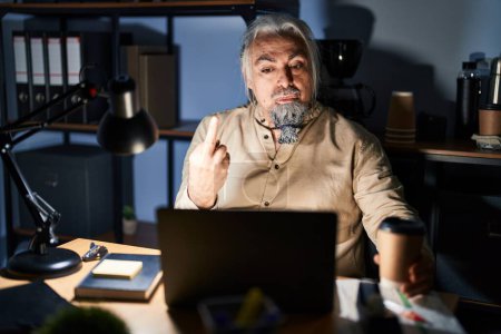 Foto de Middle age man with grey hair working at the office at night showing middle finger, impolite and rude fuck off expression - Imagen libre de derechos