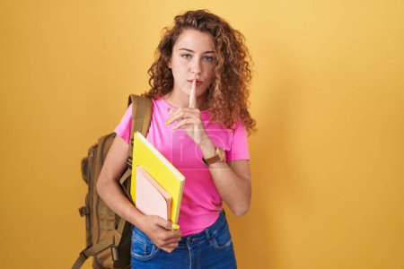 Téléchargez les photos : Young caucasian woman wearing student backpack and holding books asking to be quiet with finger on lips. silence and secret concept. - en image libre de droit