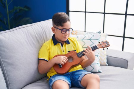 Téléchargez les photos : Adorable garçon hispanique jouant ukulele assis sur le canapé à la maison - en image libre de droit