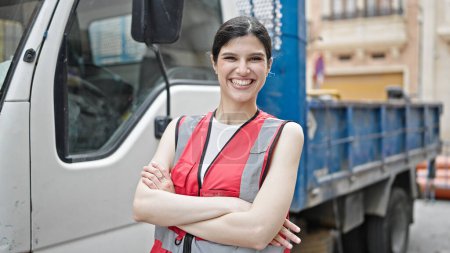 Foto de Joven hermosa mujer hispana constructora sonriendo confiada de pie con los brazos cruzados gesto por camión en la calle - Imagen libre de derechos