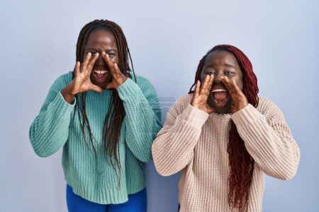 Foto de Dos mujeres africanas de pie sobre fondo azul gritando furiosas con las manos sobre la boca - Imagen libre de derechos