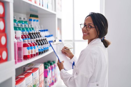 Téléchargez les photos : Jeune pharmacienne hispanique écrivant sur des étagères à la pharmacie - en image libre de droit