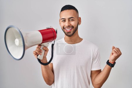 Photo for Young hispanic man shouting through megaphone screaming proud, celebrating victory and success very excited with raised arm - Royalty Free Image