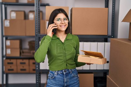 Photo for Young beautiful hispanic woman ecommerce business worker talking on smartphone holding packages at office - Royalty Free Image