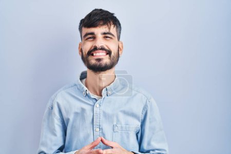 Foto de Joven hombre hispano con barba de pie sobre fondo azul manos juntas y los dedos cruzados sonriendo relajado y alegre. éxito y optimismo - Imagen libre de derechos