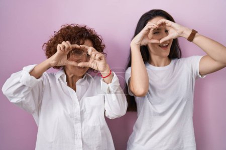 Foto de Madre e hija hispanas vistiendo camiseta blanca casual sobre fondo rosa haciendo forma de corazón con la mano y los dedos sonriendo mirando a través del signo - Imagen libre de derechos