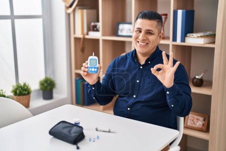 Foto de Joven hispano sosteniendo dispositivo glucosímetro haciendo signo de ok con los dedos, sonriendo gesto amistoso excelente símbolo - Imagen libre de derechos
