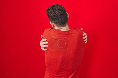 Téléchargez les photos : Jeune homme hispanique debout sur fond rouge se serrant dans ses bras heureux et positif à partir de l'arrière. amour de soi et soins de soi - en image libre de droit