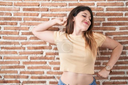 Téléchargez les photos : Jeune femme brune debout sur un mur de briques s'étirant vers l'arrière, fatiguée et détendue, endormie et bâillant tôt le matin - en image libre de droit