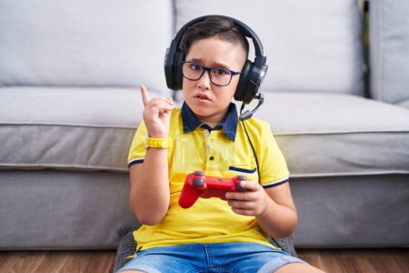 Foto de Joven niño hispano jugando videojuego sosteniendo controlador usando auriculares apuntando a un lado preocupado y nervioso con el dedo índice, preocupado y sorprendido expresión - Imagen libre de derechos