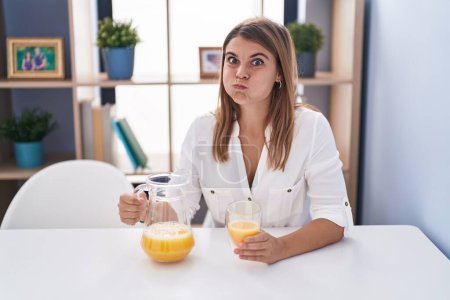 Téléchargez les photos : Jeune femme hispanique buvant un verre de jus d'orange gonflant les joues avec un visage drôle. bouche gonflée d'air, attrapant l'air. - en image libre de droit