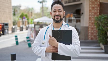 Téléchargez les photos : Jeune homme latin médecin souriant confiant tenant rapport médical à la terrasse du café - en image libre de droit
