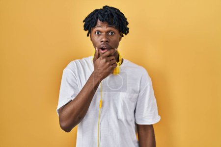 Photo for Young african man with dreadlocks standing over yellow background looking fascinated with disbelief, surprise and amazed expression with hands on chin - Royalty Free Image