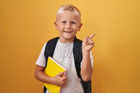 Foto de Pequeño chico caucásico usando mochila de estudiante y sosteniendo el libro sonriendo feliz señalando con la mano y el dedo hacia un lado - Imagen libre de derechos